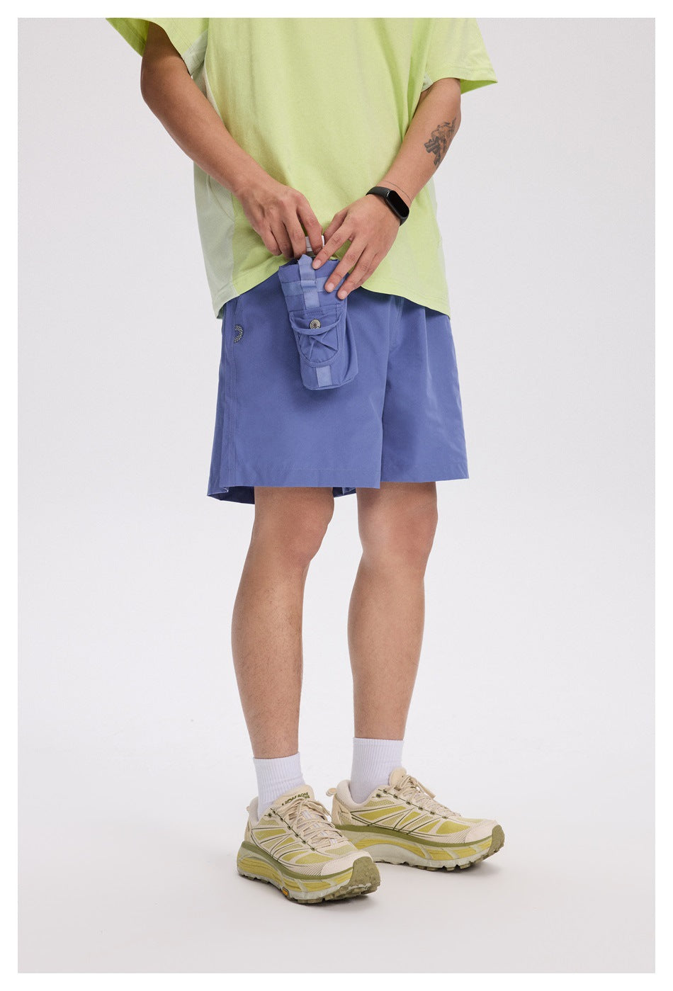 Model wearing periwinkle-blue shorts and holding a matching utility pouch, with a light lime T-shirt and beige sneakers.