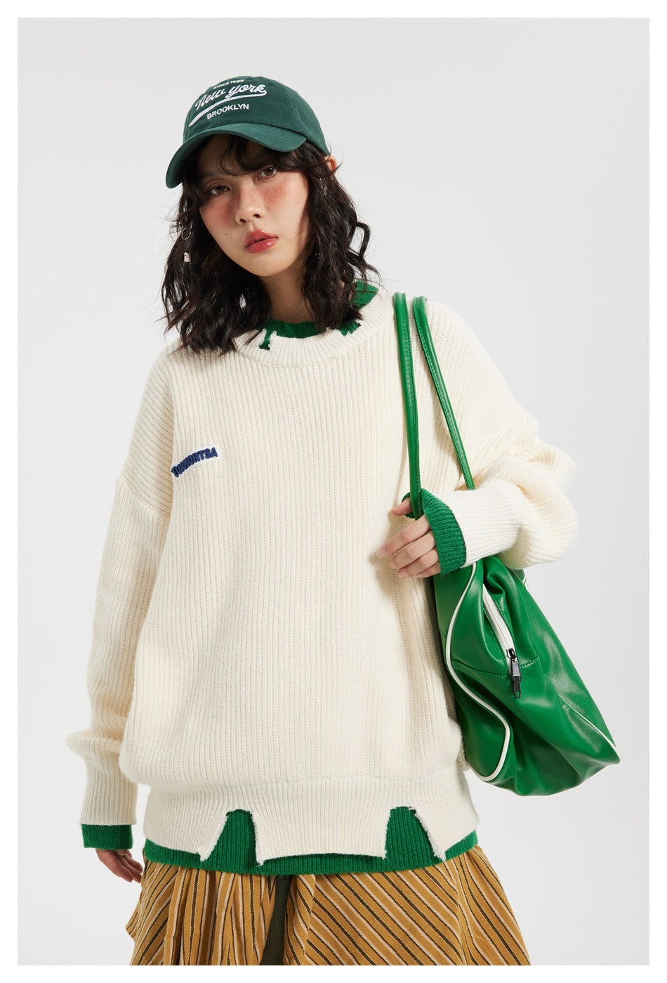 Model wearing an ivory sweater with green trim and a scalloped hem, shown from the front.
