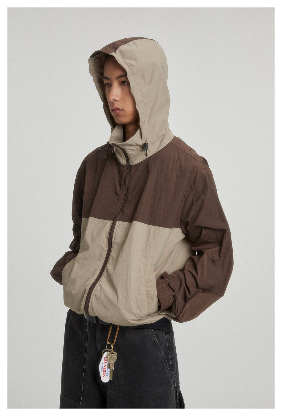 Model wearing brown and khaki hooded zip jacket with hood up, hand in pocket, with dark jeans and a keychain