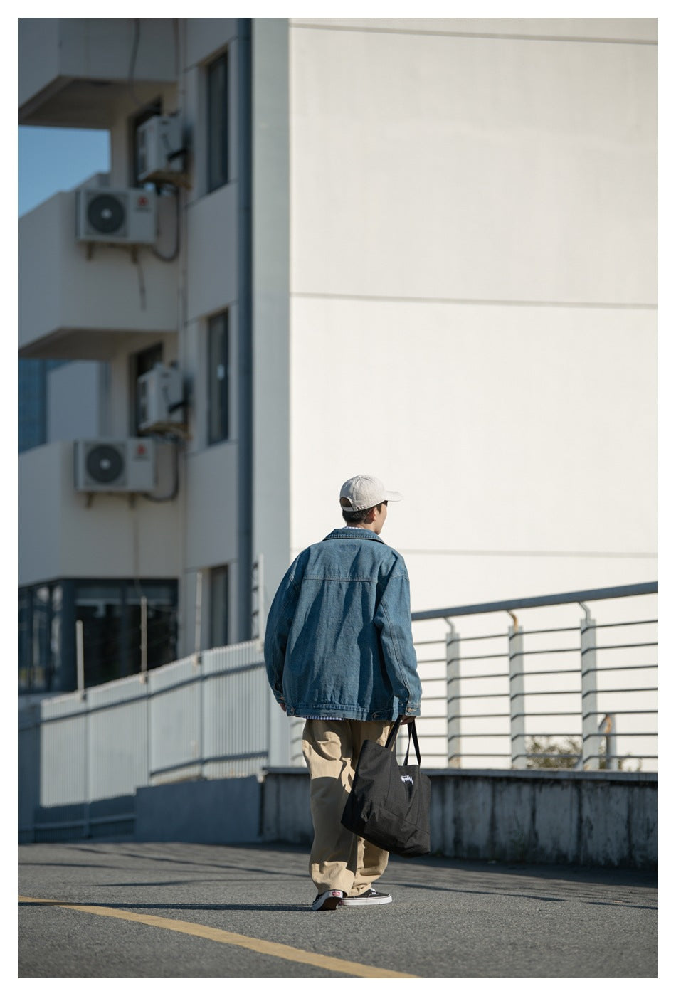 Back view of a model walking away in a blue denim jacket, cap, khaki pants and a black tote bag