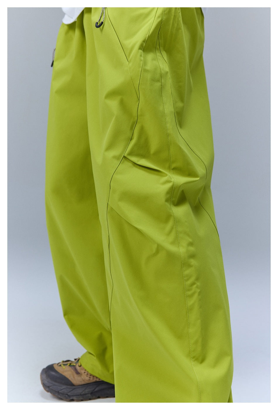 Close side view of a person wearing bright lime-green loose pants with curved seam panels and a hiking shoe.