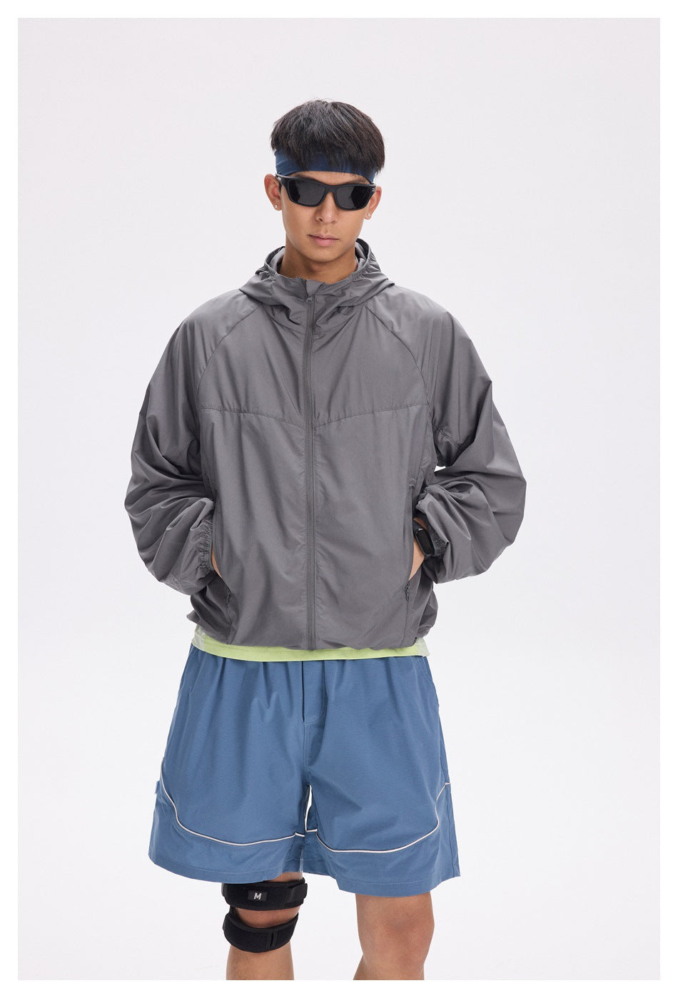 Front view of a model in a charcoal gray hooded zip jacket and blue shorts, with sunglasses and a blue headband.