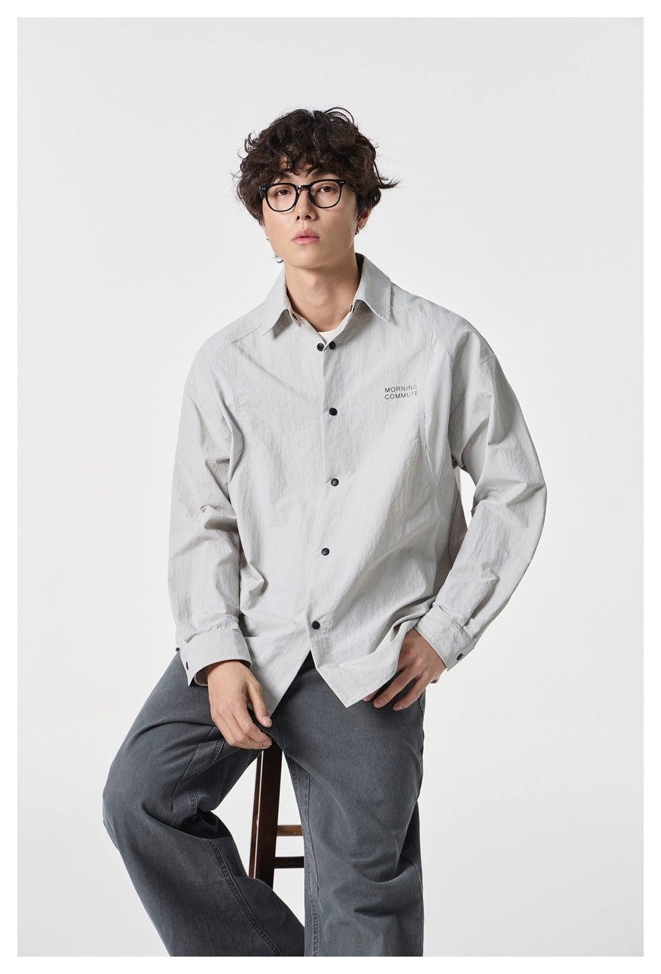 Man in glasses seated on a stool wearing a light gray button-front shirt with gray pants