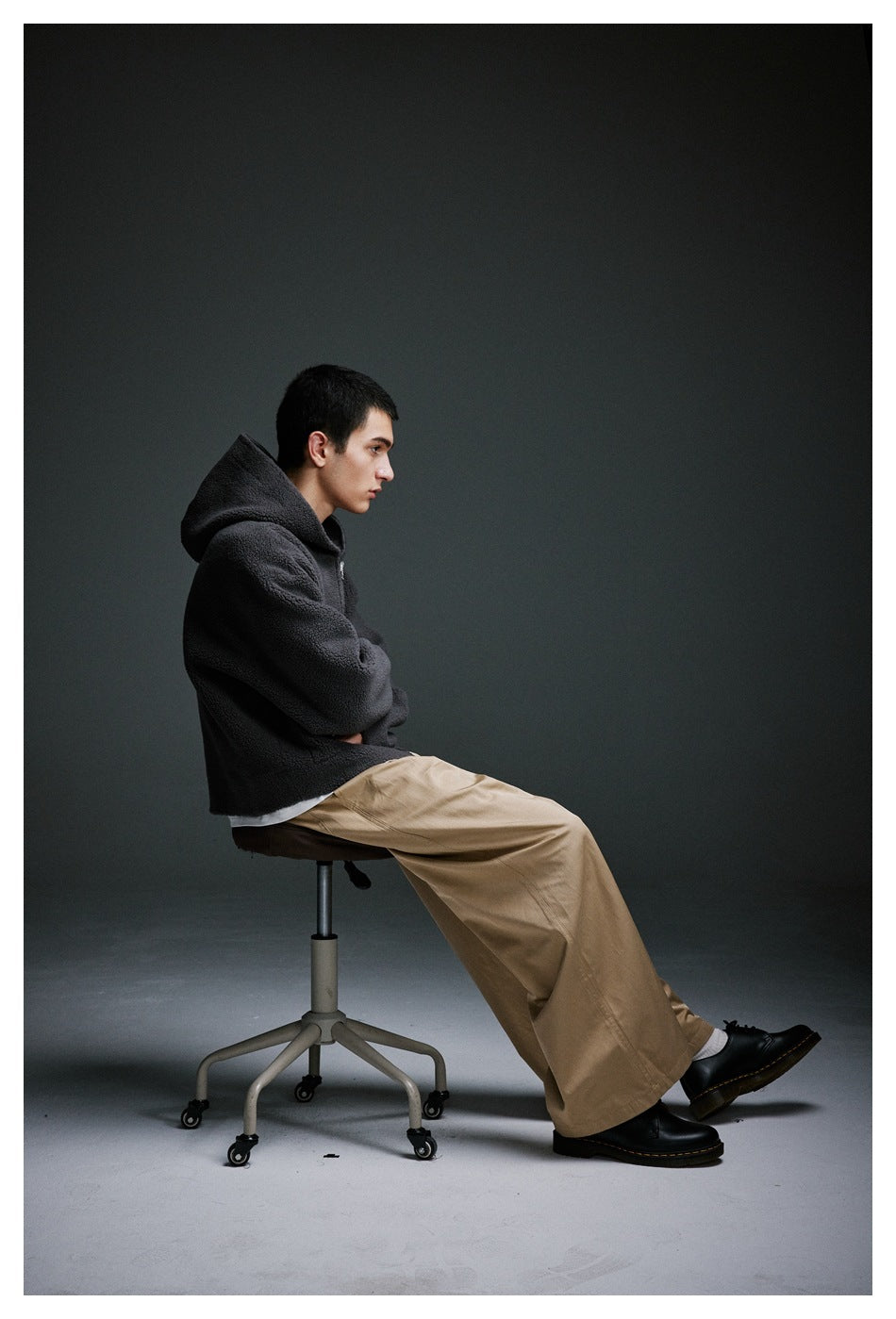 Seated side view of a model in a charcoal hooded jacket and wide taupe trousers.