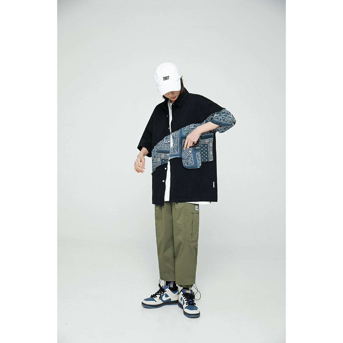 Model wearing an oversized black shirt with a blue bandana-print panel, olive cargo pants, and a white cap.