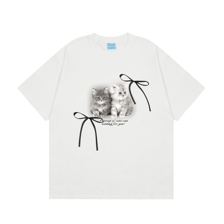 White short-sleeve crew-neck T-shirt with a kitten graphic, two black ribbon bows and script text on the front