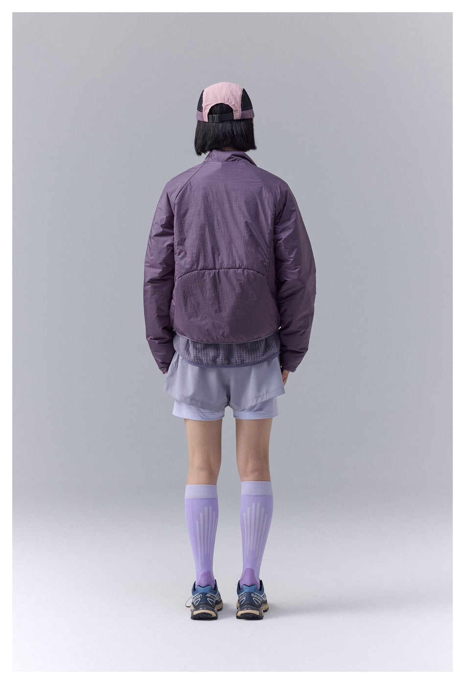 Rear view of a model wearing a purple jacket, cap, pale shorts, and knee-high lavender socks.