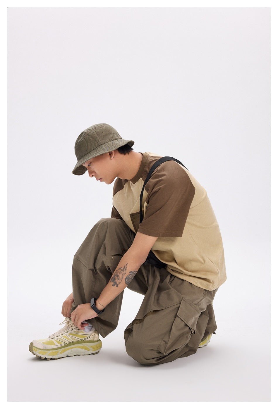 Model crouching in a beige and brown color-block T-shirt, olive cargo pants, and a khaki bucket hat.