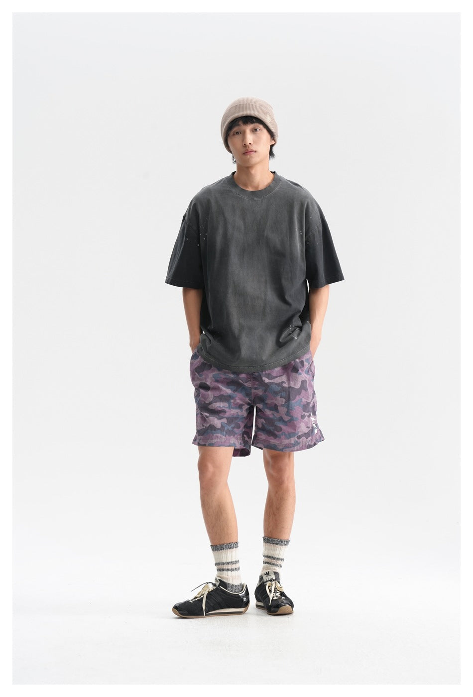 Full-length front view of a model wearing a charcoal oversized T-shirt, purple camouflage shorts, and a beige knit beanie.