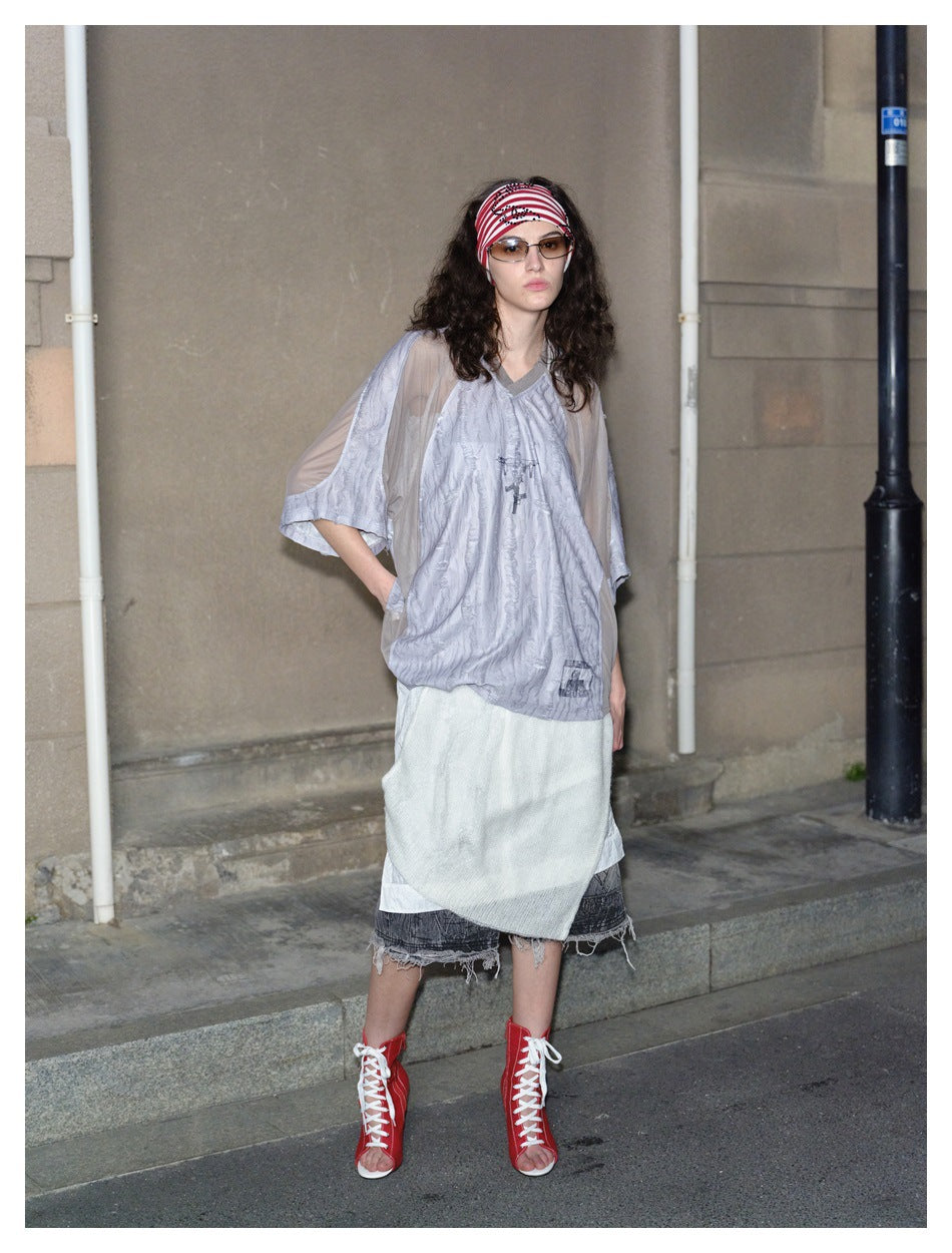 Full-length model wearing a pale gray loose top, layered white and dark skirt, and red lace-up shoes.