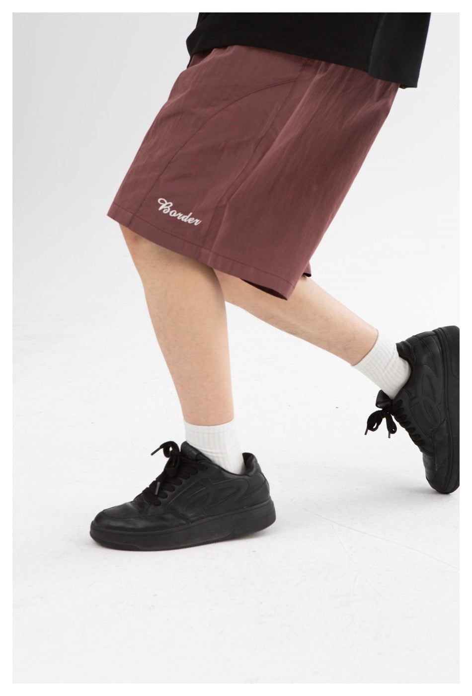 Close view of a model wearing mauve shorts with white script embroidery, white socks, and black sneakers.