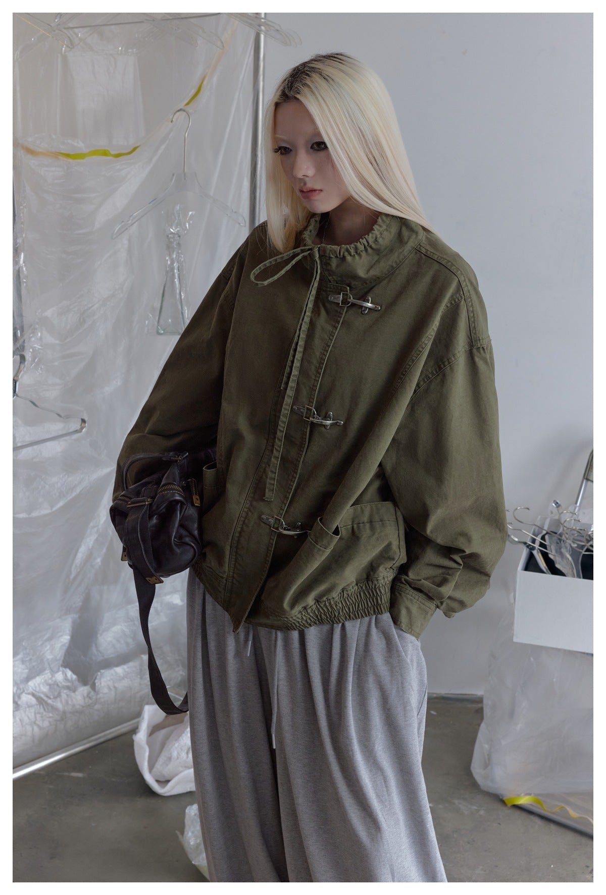Model wearing an oversized olive jacket with a high gathered collar, metal hook closures and elasticated hem.