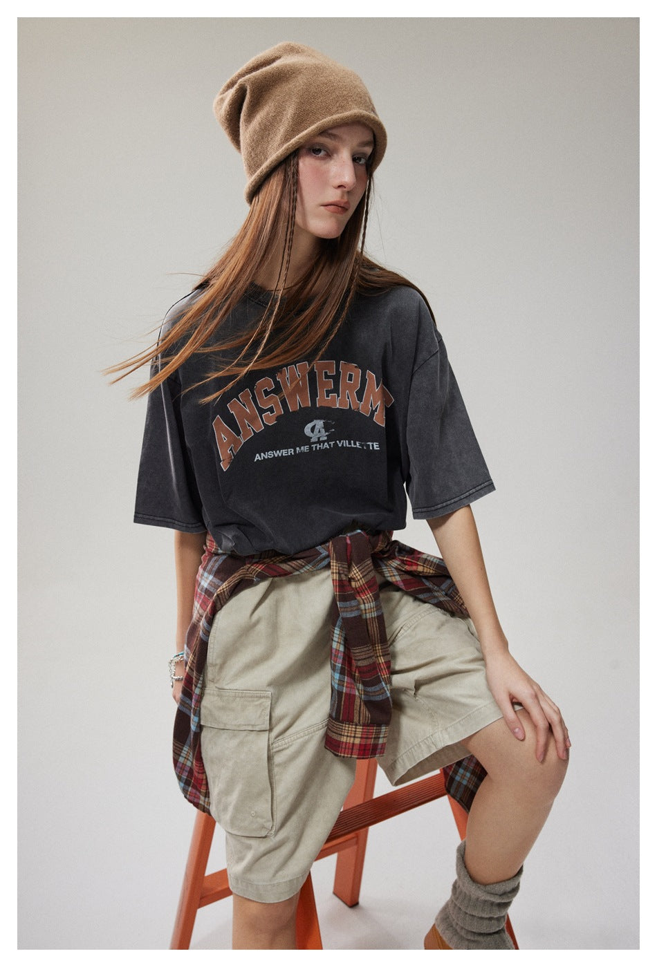 Seated model wearing a charcoal graphic T-shirt, beige shorts, a tan beanie, and a plaid shirt tied at the waist.