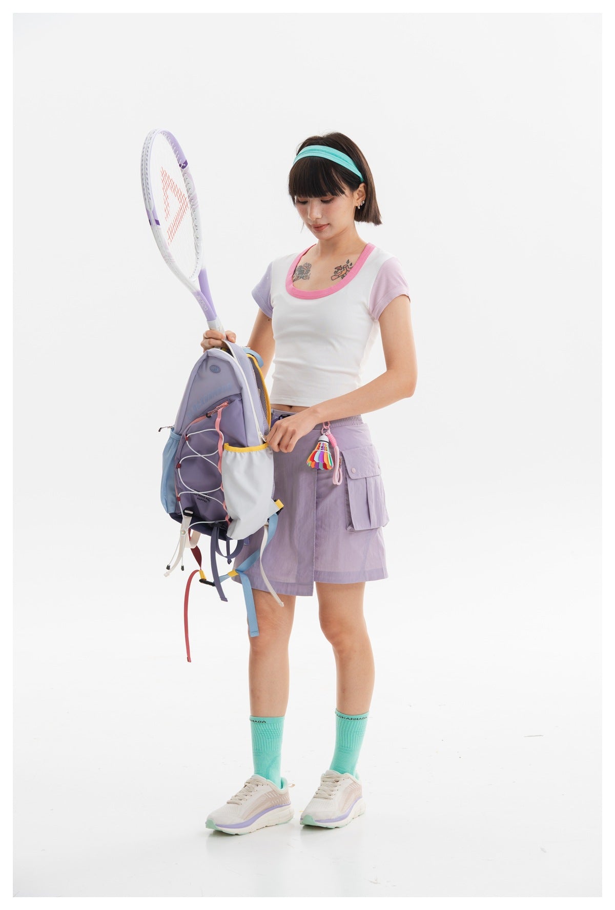 Model in a white, pink-trimmed crop top and lavender cargo skirt, holding a tennis racket beside a backpack.