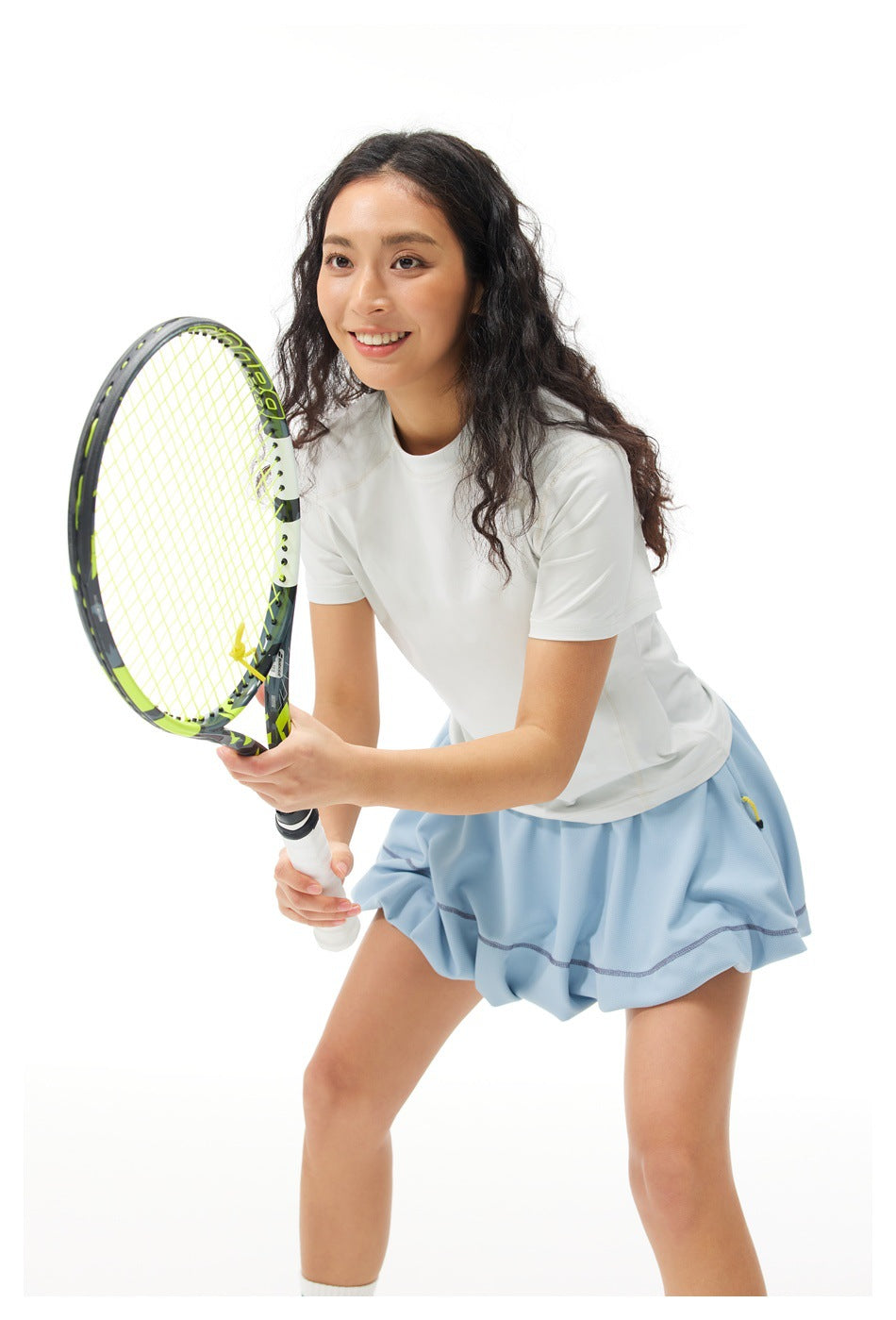 Model wearing a white short-sleeve top and pale blue tennis skirt, holding a racket in a ready stance.