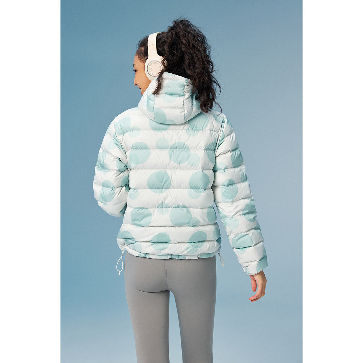 Model viewed from behind wearing a white hooded puffer jacket with large pale aqua circles and gray leggings.