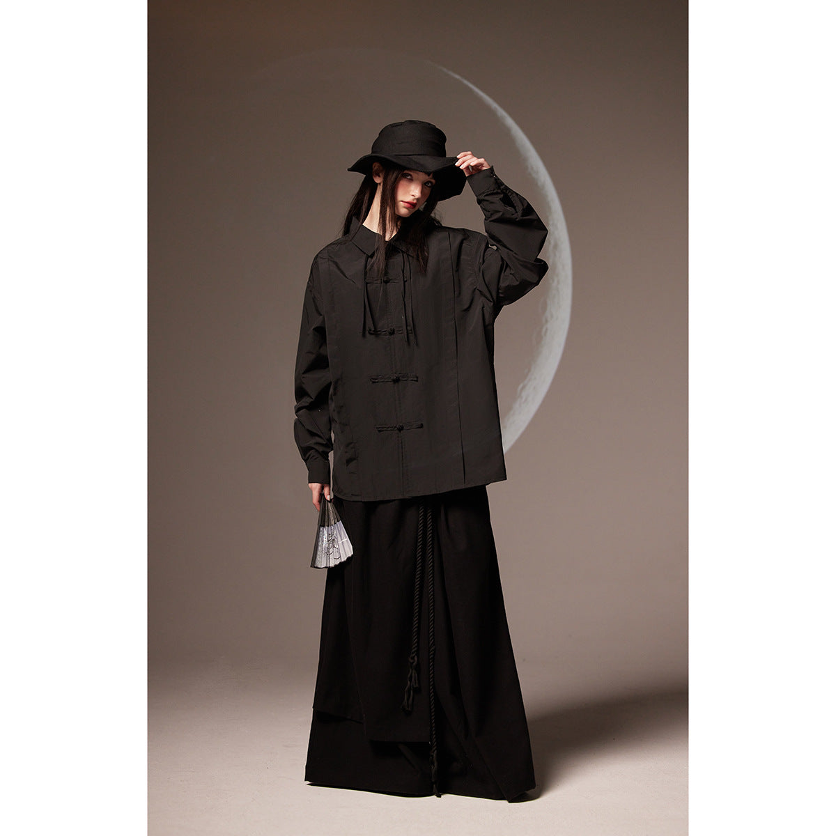Model wearing a black oversized shirt with frog closures, a long black skirt, and a black bucket hat.