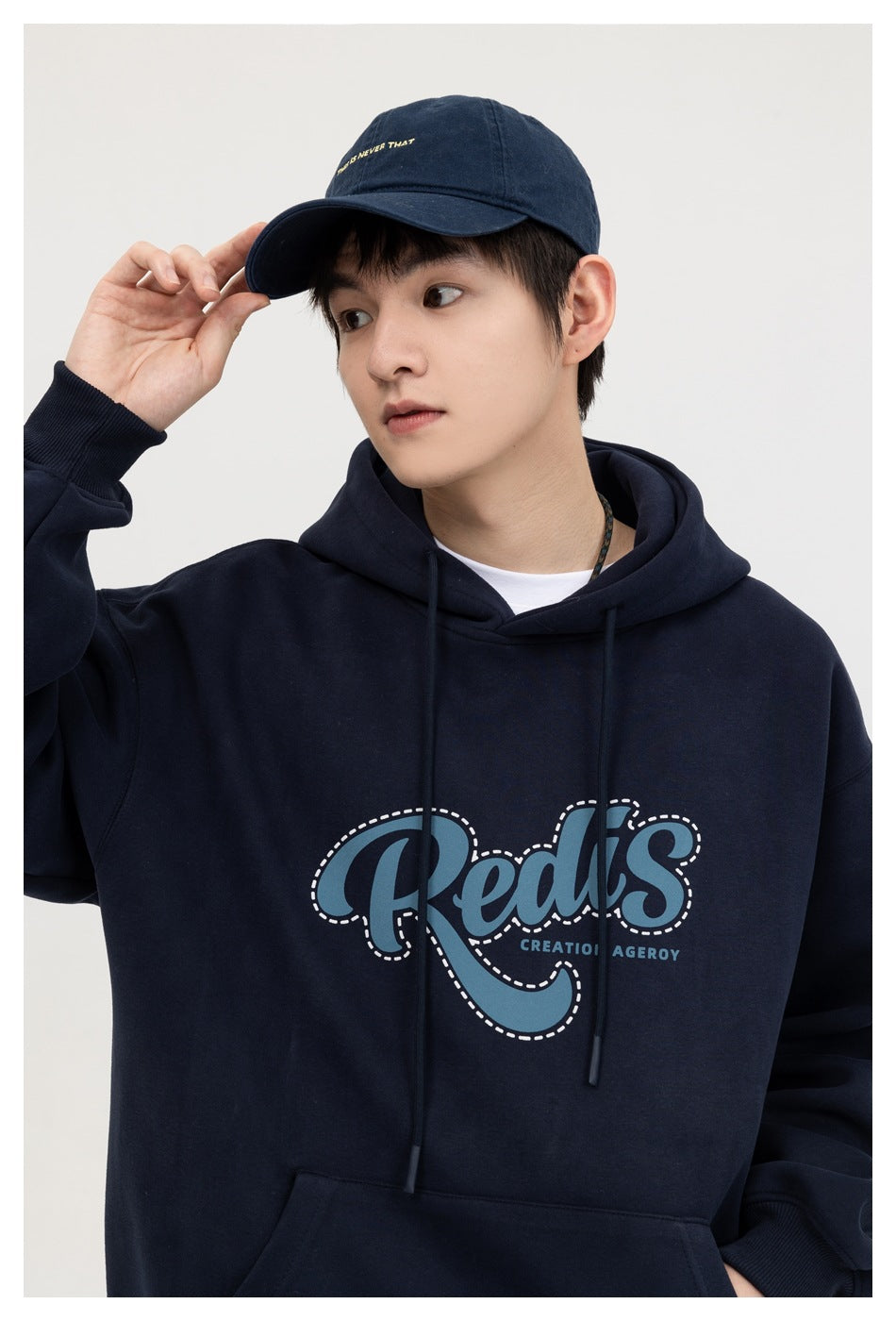 Model in a navy graphic hoodie and matching cap, shown at an angle while touching the cap brim.