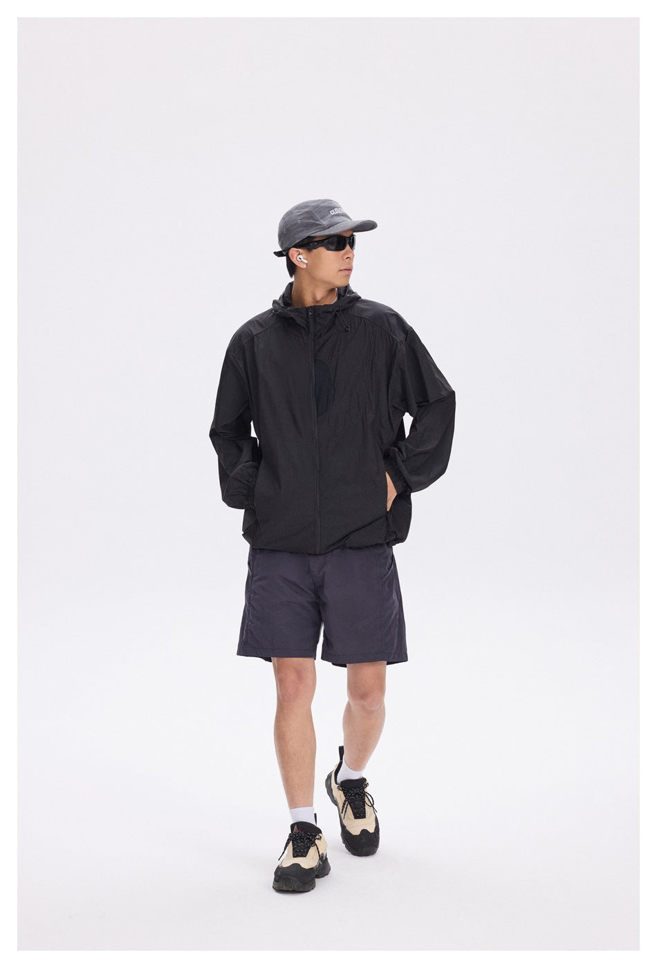 Model wearing a black hooded zip jacket with gray cap, sunglasses, navy shorts and sneakers