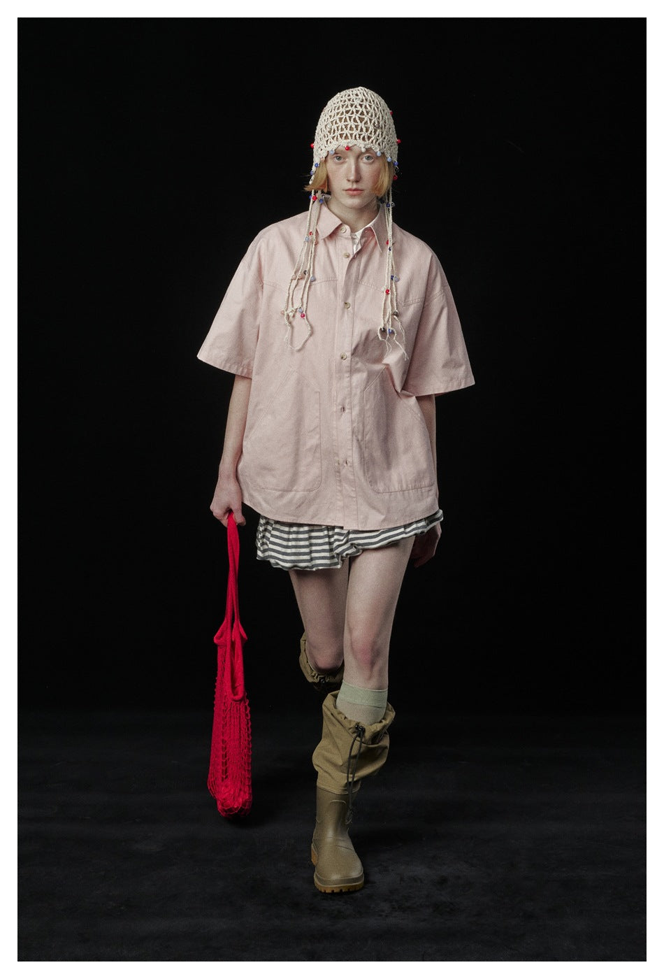 Front view of a model wearing an oversized pale pink button-up shirt, striped shorts, and a beaded crochet cap.