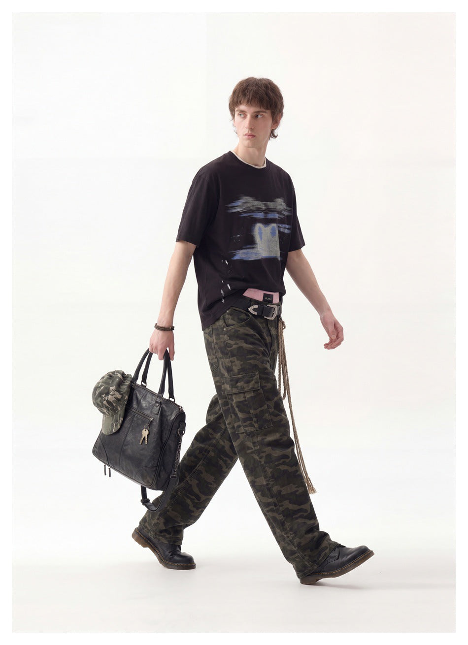 Model walking in a black graphic T-shirt and loose camouflage trousers, carrying a black bag.