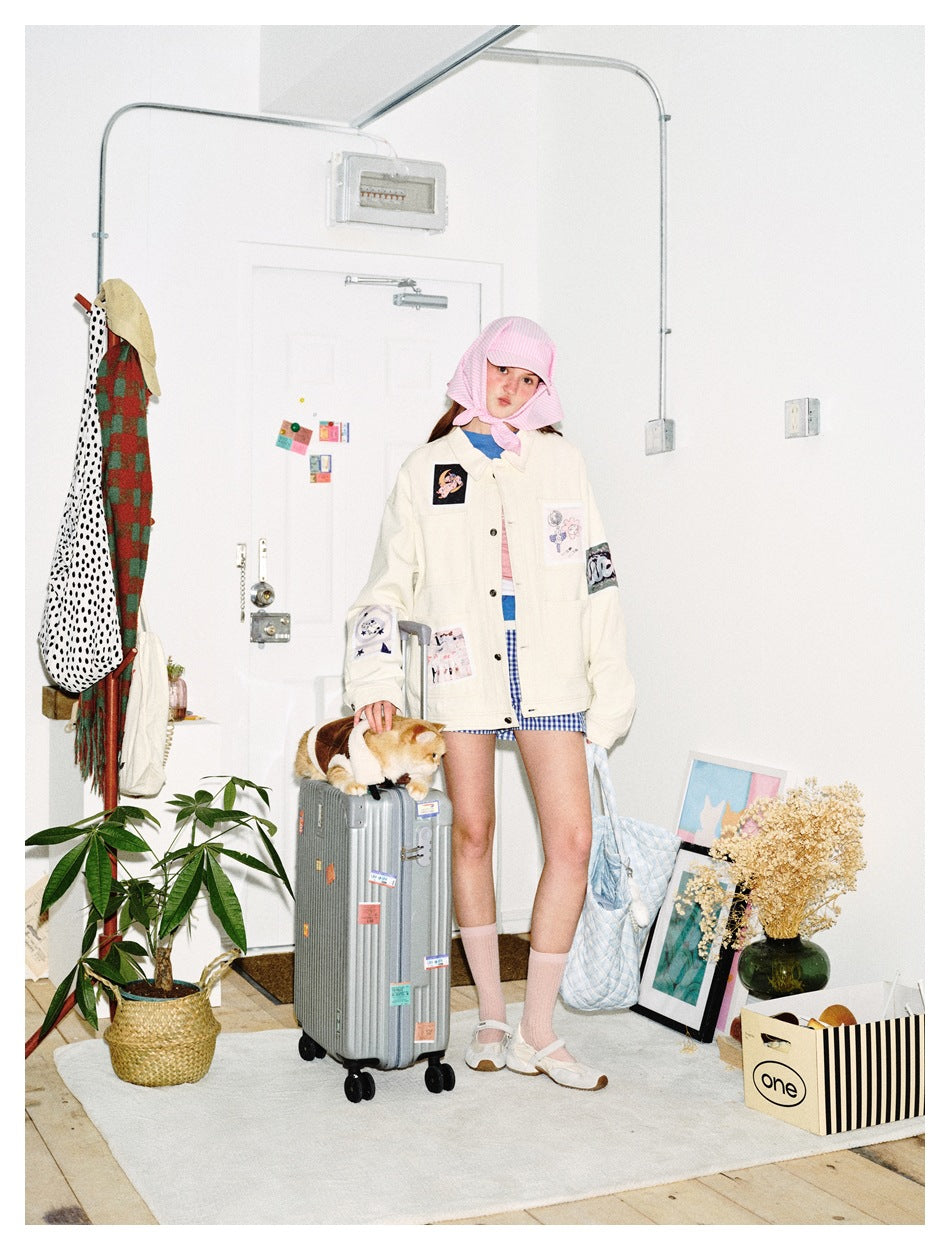 Model in a beige patch-detail jacket, pink headscarf and gingham shorts, standing beside a suitcase with a cat on it
