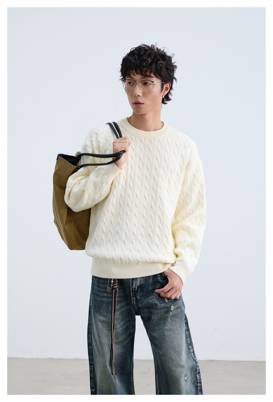 Model wearing an ivory cable-knit crewneck sweater and dark jeans, carrying a tan tote bag.