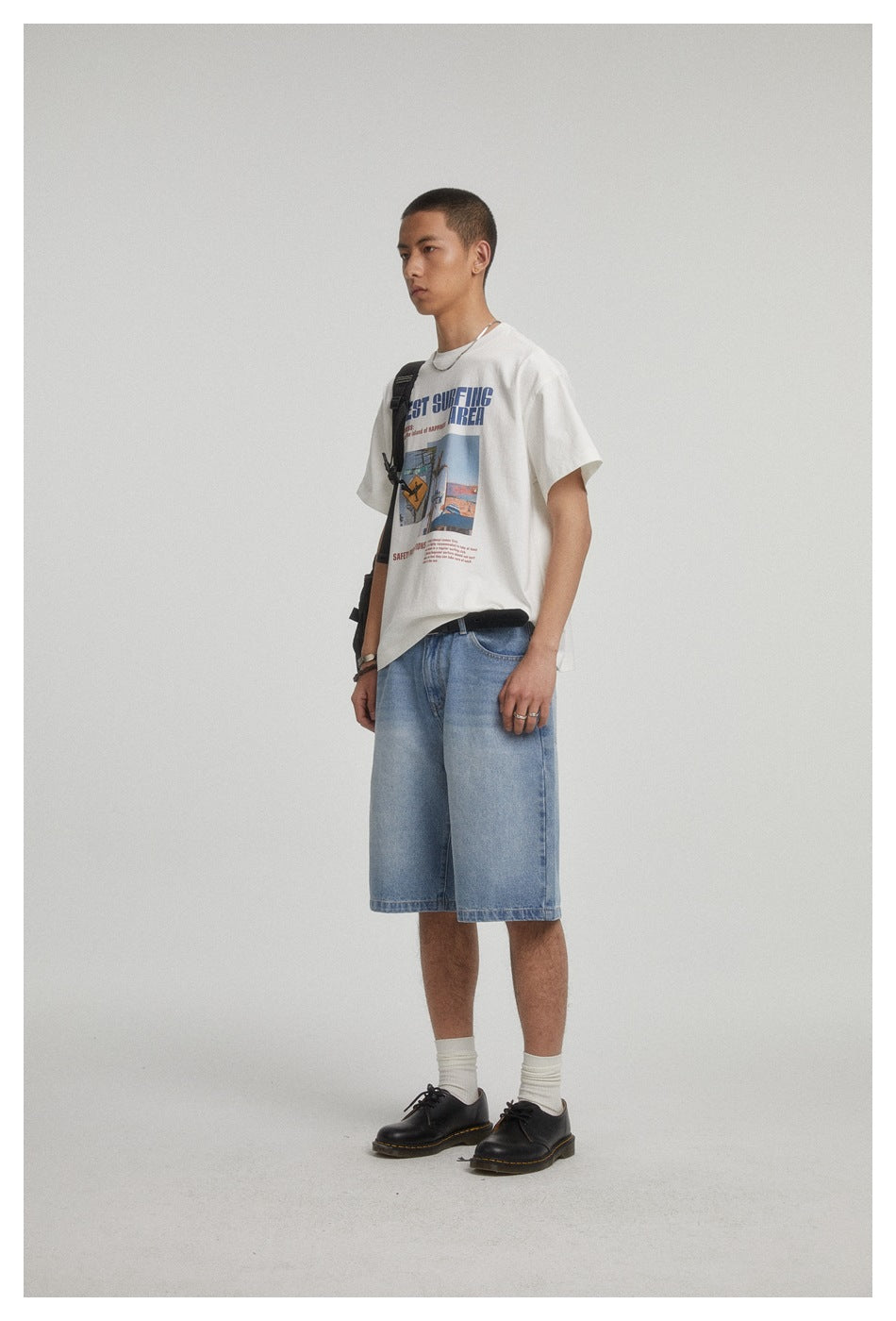 Side view of a model wearing a white graphic T-shirt, light blue denim shorts, and a black shoulder bag.