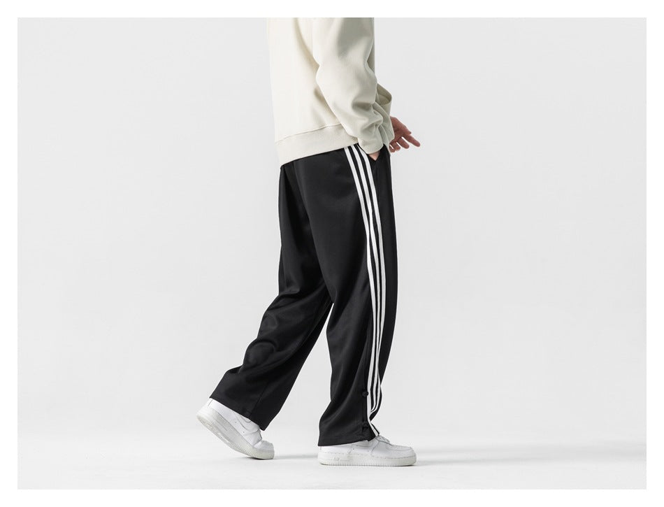Rear three-quarter view of a model wearing loose black track pants with three white side stripes.