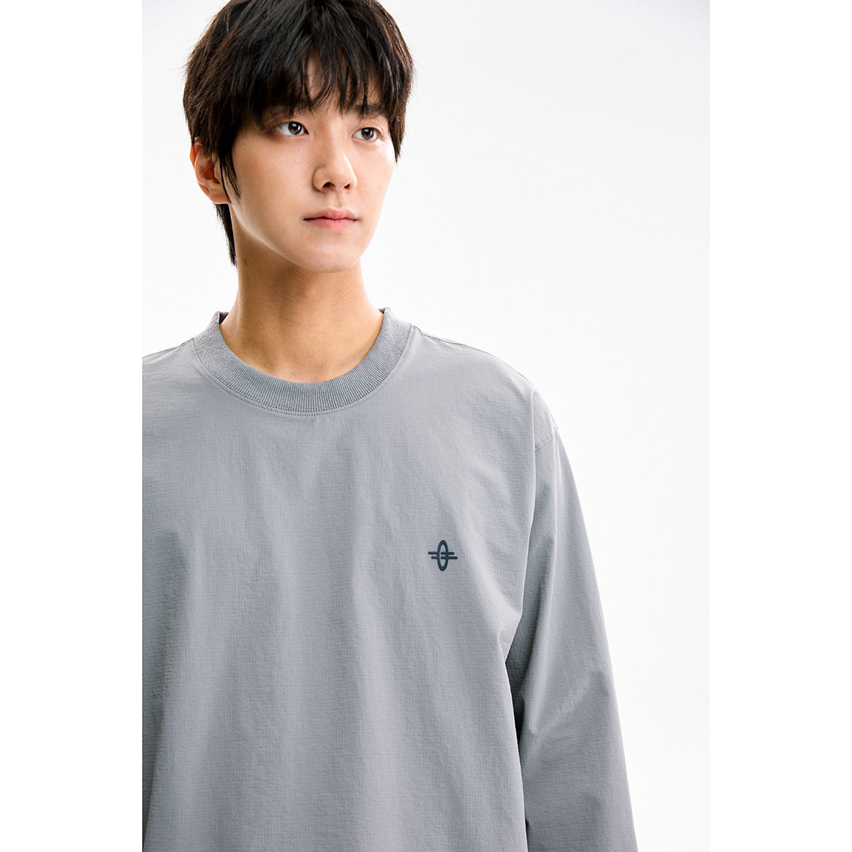 Close-up of a model wearing a gray crewneck shirt with ribbed collar and a small dark chest emblem.