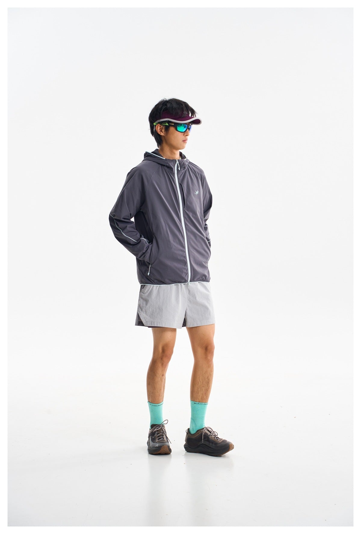 Model wearing a charcoal hooded jacket with pale piping and a front zipper, paired with light gray shorts.