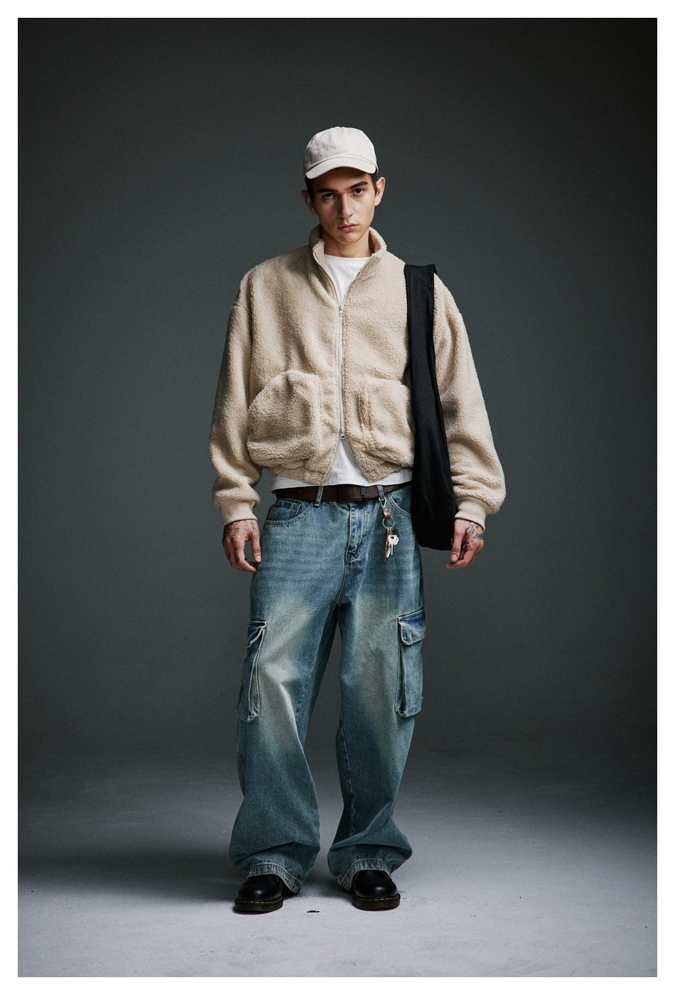 Full-length model wearing a cream zip-up jacket, white cap, oversized blue cargo jeans and a black shoulder bag.
