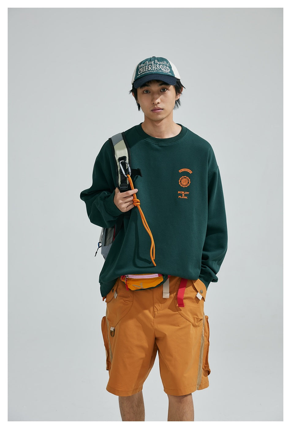 Closer view of a model in a dark green crewneck sweatshirt with a small orange chest graphic and mustard cargo shorts.