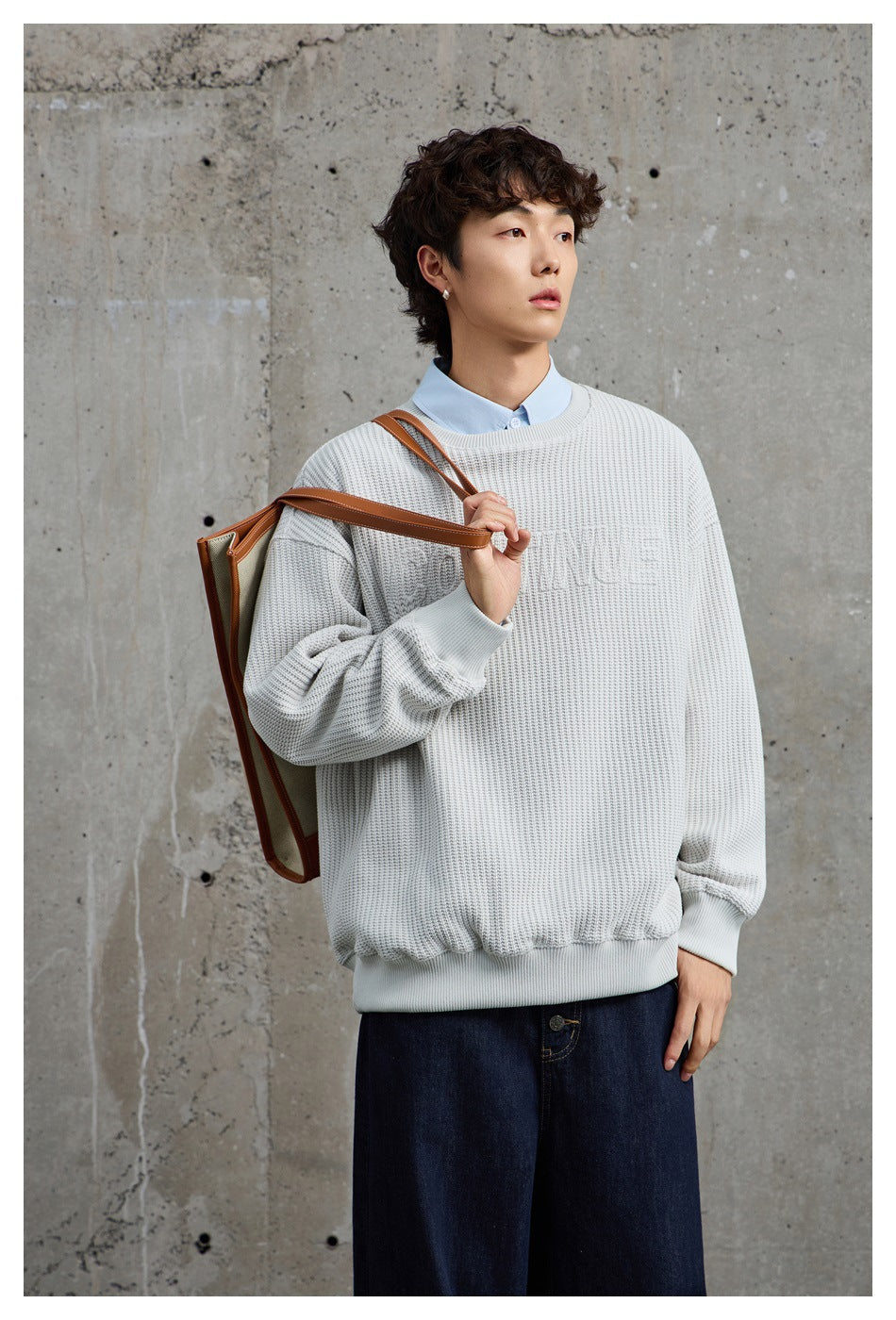 Model wearing a light gray textured crewneck sweatshirt over a blue collared shirt, holding a tote bag on his shoulder