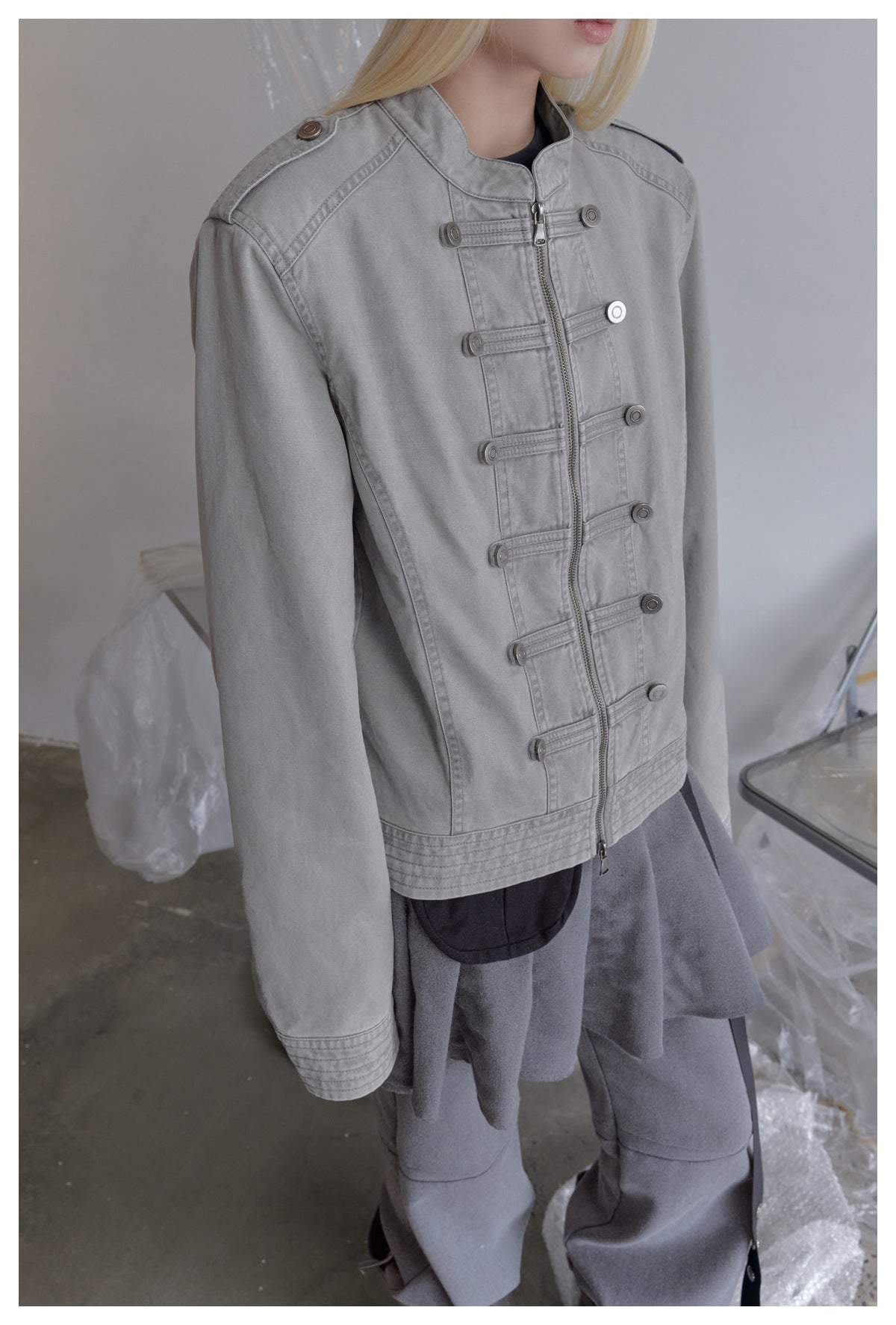 Close view of a light gray jacket showing its front zipper, horizontal button tabs, shoulder epaulette, and stitched cuffs.