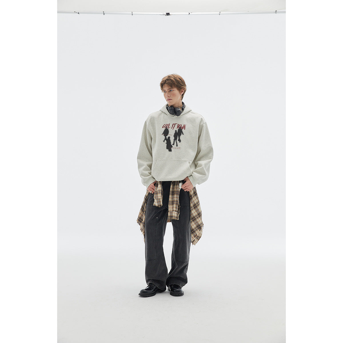 Model wearing a light gray graphic hoodie with a kangaroo pocket, plaid shirt tied at the waist, and dark wide-leg pants.