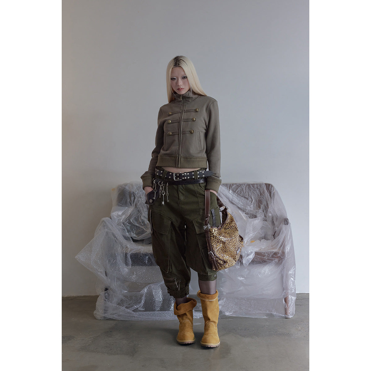 Model wearing a taupe cropped zip jacket with brass button tabs, olive cargo pants, and tan boots.