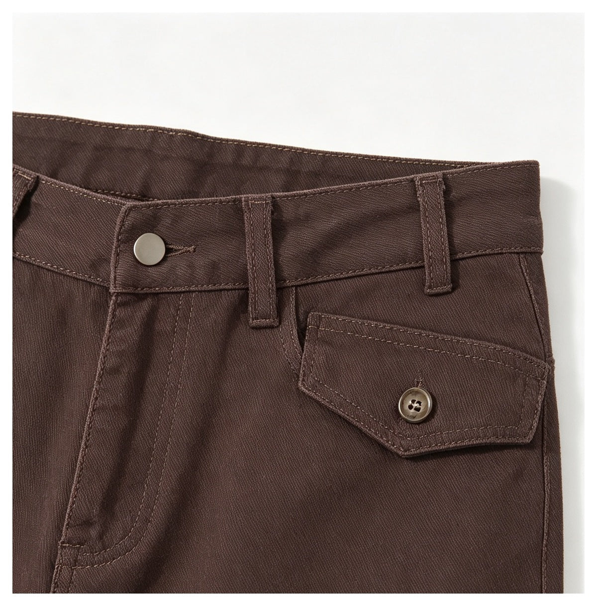 Close-up of the brown trousers' waistband, belt loops, front pocket opening, and buttoned flap pocket.