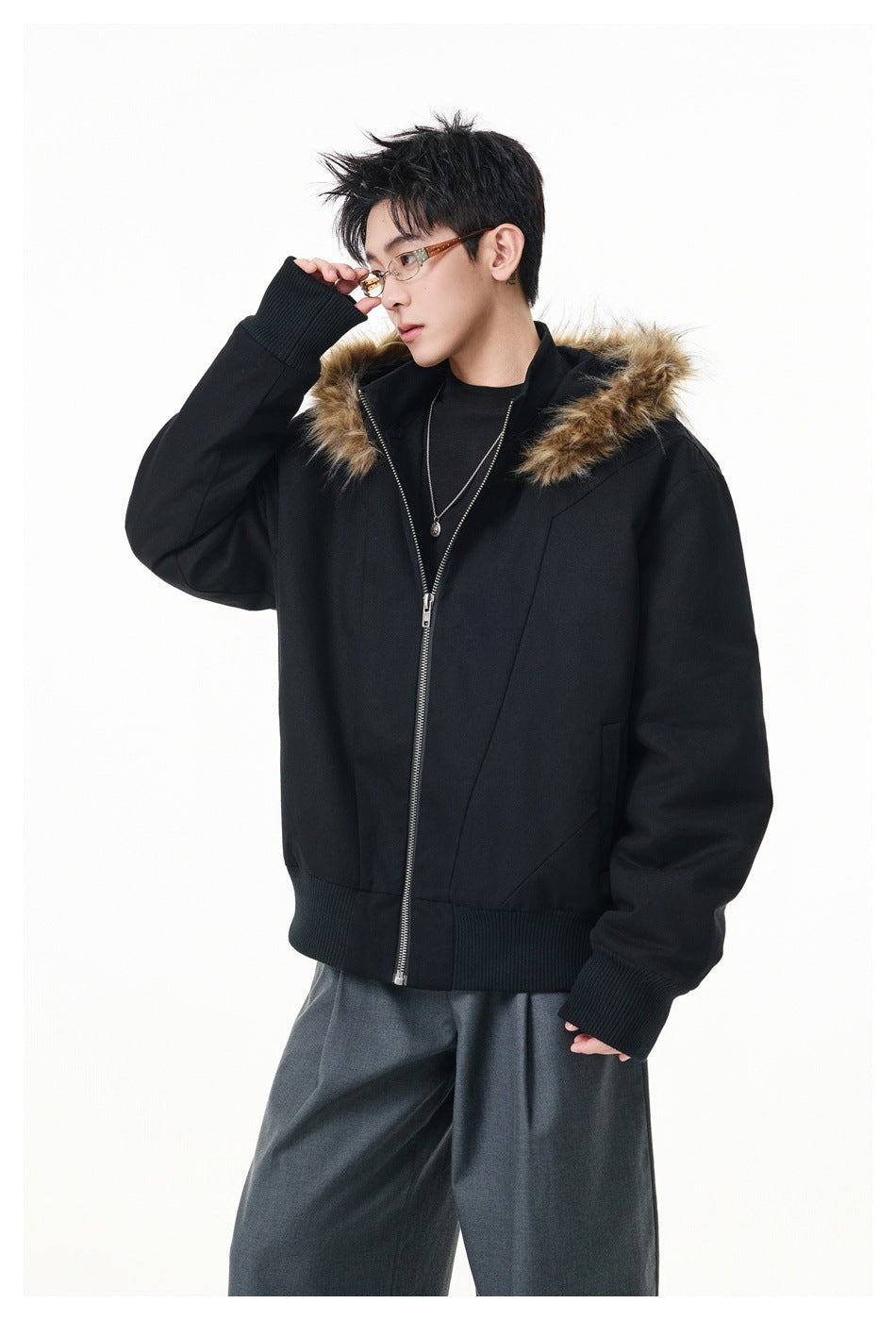 Model wearing a black zip jacket with a hood trimmed in fur-like brown fibers, ribbed cuffs and hem, with gray trousers