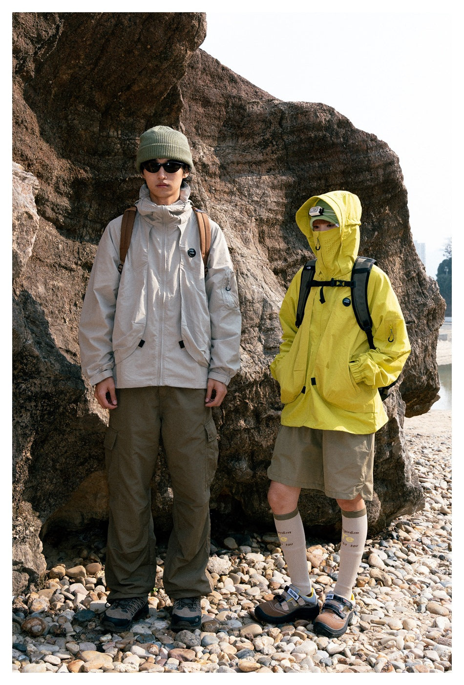 Two models outdoors wearing a light beige and a bright yellow hooded jacket with cargo bottoms.