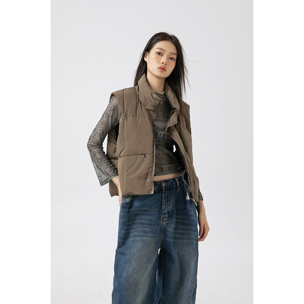 Model wearing an open taupe padded vest over a gray patterned sheer top and wide-leg blue jeans.