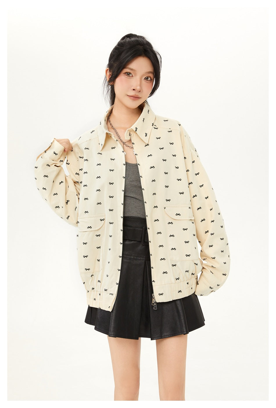 Model wearing a cream jacket with small black bow motifs and flap pockets, layered over a gray top and black skirt.