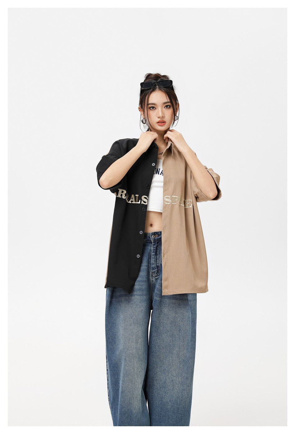 Model adjusts the collar of an oversized black-and-tan embroidered shirt worn open over a white crop top and jeans.