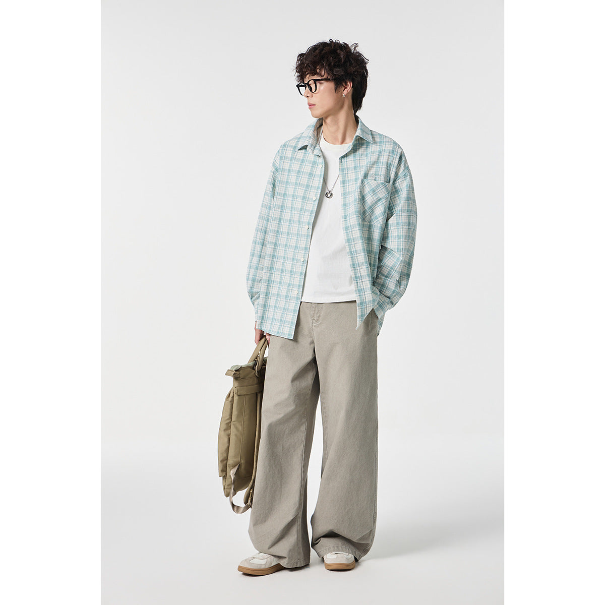 Model in a pale blue plaid shirt and loose taupe trousers, carrying an olive bag.