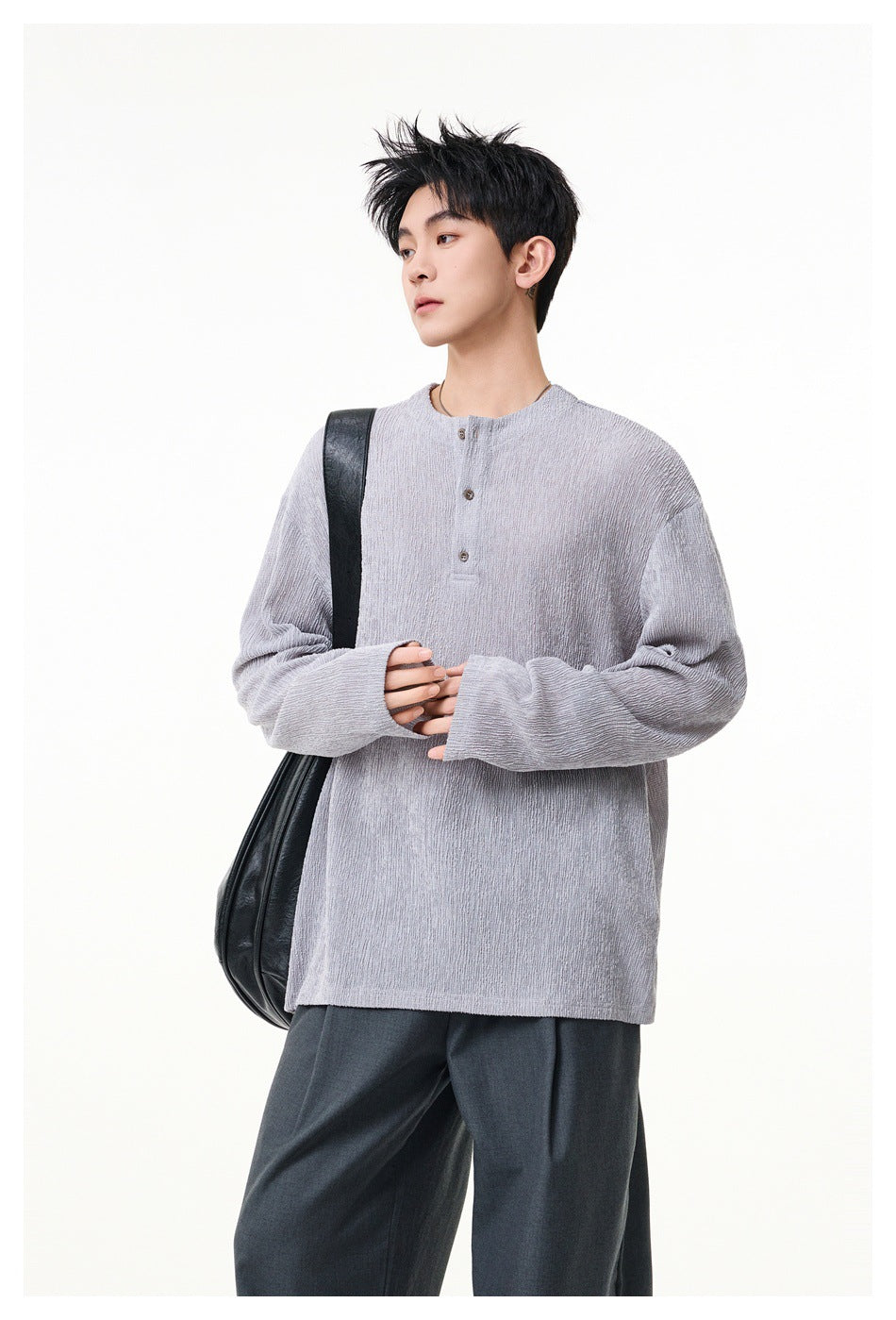 Model wearing a light gray oversized henley with a short button placket and dark trousers, carrying a black shoulder bag.
