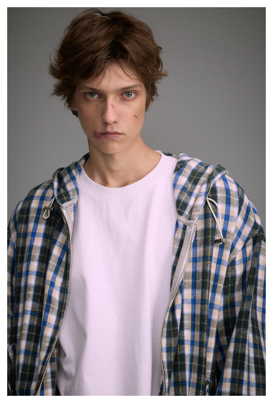 Close-up of a model in a blue plaid hooded jacket, open zipper and drawstring toggles over a white tee