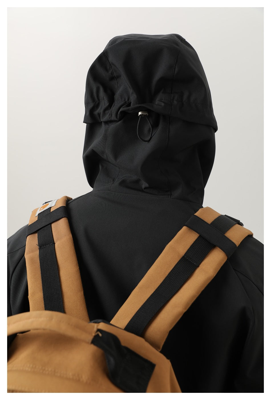 Rear view of a model wearing a black hooded jacket, with the hood and backpack straps visible.