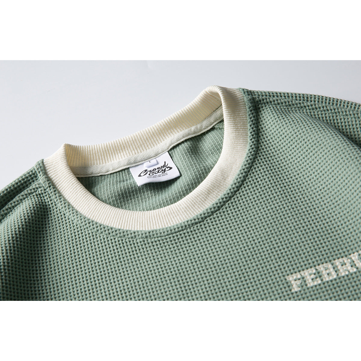 Close-up of a sage-green textured sweatshirt neckline with an ivory ribbed collar and sewn neck label.