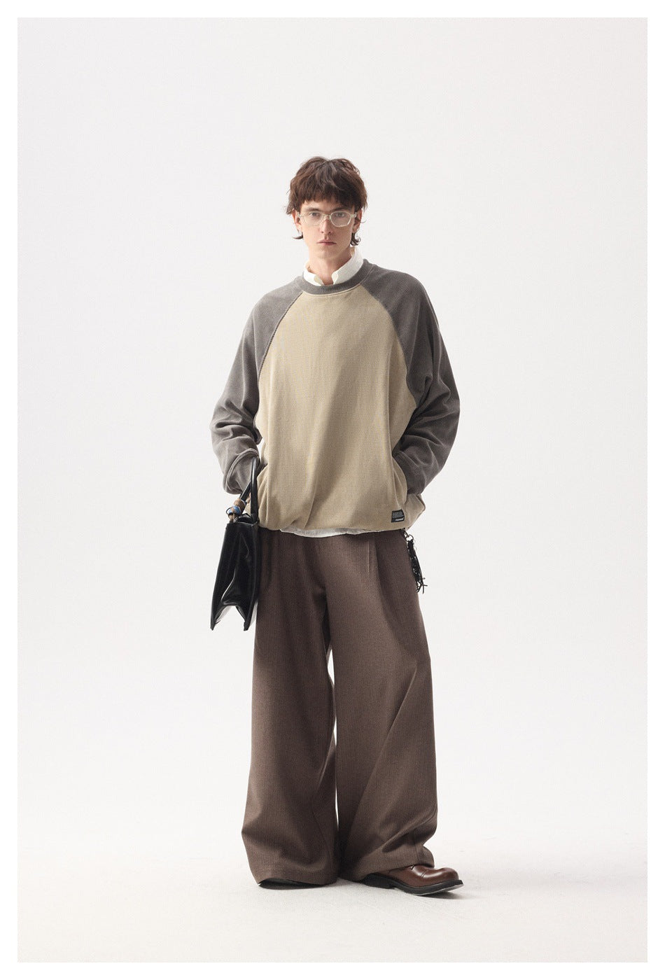 Full-length view of a model in a tan and taupe raglan sweatshirt with wide brown trousers and a black bag
