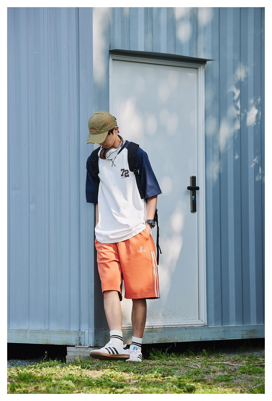 Model leaning against a door in the white and navy raglan T-shirt with orange shorts, cap, headphones and backpack